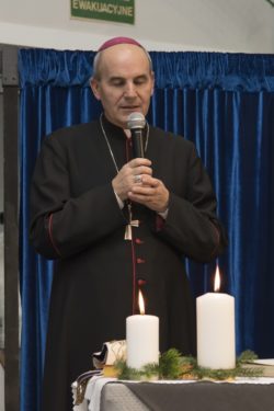 Biskup mówi do mikrofonu. Przednim udekorowany stół z zapalonymi świecami.