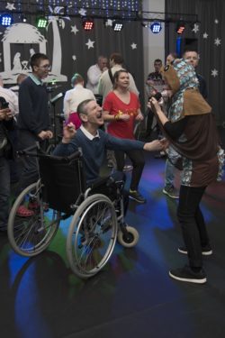 Uśmiechnięty mężczyzna na wózku inwalidzkim tańczy z osobą w zabawnym przebraniu.