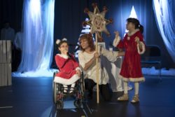 Od lewej: dziewczynak na wózku inwalidzkim z mikrofonem w dłoni. Obok niej kobieta trzyma gwiazdę betlejemską. obok kobiety stoi dziewczynka.