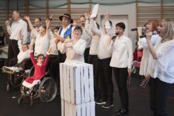 Na sali gimnastycznej stoi grupa odświętnie ubranych osób z mikrofonami w dłoniach, w tym dwie na wózkach inwalidzkich. Pięć z nich unosi ręce do góry.