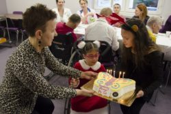 Kobieta i dziewczynka trzymają tort przed uśmiechniętą dziewczynką na wózku inwalidzkim.