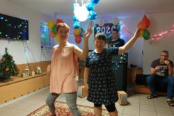 Dwie mieszkanki tańczą na tle dekoracji sylwestrowych - kolorowe balony, wstąrzki, lampki.
