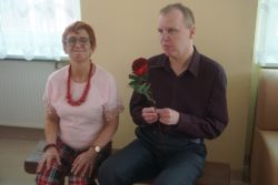 Na ławeczce siedzi kobieta i mężczyzna. Mężczyzna trzyma w rękach czerwoną różę.