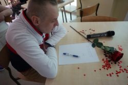 Przy stoliku na krześle siedzi mężczyzna. Na stoliku leży czerwona róża, kartka i na kartce długopis.
