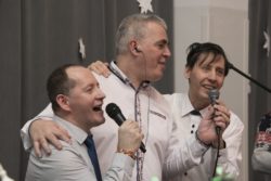 Trzech mężczyzn w jasnych koszulach śpiewa do mikrofonów.