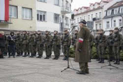 Starszy mężczyzna w mundurze stoi przed mikrofonem. W tle stoją umundurowani żołnierze.