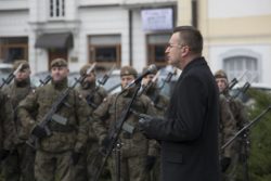 Mężczyzna stoi przed mikrofonem. W tle żołnierze.