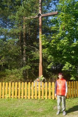 Kobieta stoi przed płotkiem za którym jest duży drewniany krzyż