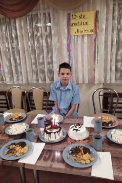 Za zasatwinym słodyczami stołem na ktrym stoi tort siedzi chłopiec. Nad chłopcem napis 15-ste urodziny Andrzeja