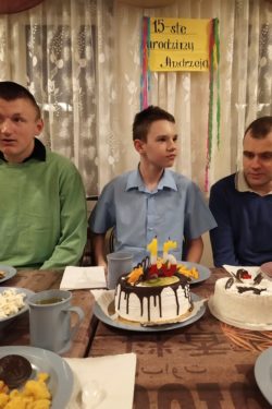 Za zastawionym słodyczami stołem na którym stoi tort siedzą trzej chłopcy. Nad sirodkowym wisi napis 15-ste urodziny Andrzeja