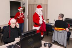 Duża sala. Na przeciw monitorów komputerowych siedzi dwóch mężczyzn. Pośrodku sali stoi Mikołaj. W drzwiach do sali stoi pani w czerwonej czapce z warkoczykami.