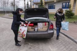 Dwie kobiety trzymające w rękach torby stoją po obu stronach samochodu z otwartym bagarznikiem. W bagażniku pudła z prezentami.