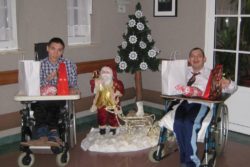 W pomieszczeniu dwóch chłopców na wózkach inwalidzkich przy choince. Na stolikach stoją torby z prezentami