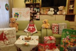 Uśmiechnięta dziewczynka w czerwonej sukience siedzi na kanapie. Wokół niej pudełka z prezentami, zabawki i stojący na stoliku tort.