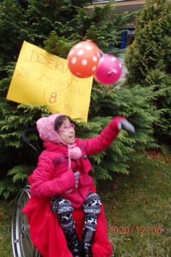 Dziewczynka na wózku inwalidzkim ubrana w różową kurtkę wypuszcza dwa baloniki. Za nią na drzewie wisi kartka z napisem: dziękuję za życzenia.