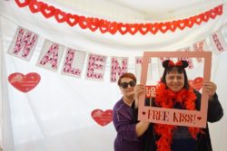 Kobieta na wysokości twarzy trzyma ramkę z napisem: free kiss! Obok niej stoi druga kobieta. W tle za nimi napis: Walentynki.