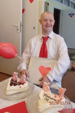 Uśmiechnięty chłopiec trzyma czerwony balonik i zapakowany prezent. Przed nim na stole stoją dwa torty w kształcie serca.