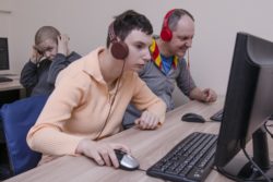 Jedna kobieta i dwóch mężczyzn z słuchawkami na uszach siedzą przed monitorami komputerowymi. 