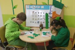 Dwie dziewczynki siedzą na krzesłach przy stoliku i przyklejają zieloną bibułkę do kartek na których narysowana jest koniczyna.