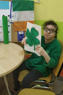 Dziewczynka siedząca na krześle przy stoliku trzyma w rękach wyklejoną na kartce zieloną koniczynę.