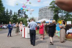 Grupa osób stoi na placu wokół dużego pudła z któreko wylatóją kolorowe balony. W tle wysoki budynek.