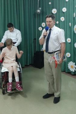 Mężczyzna z mikrofonem w ręku stoi w przyozdobionej na zielono sali. Obok stoi chłopiec i dziewczynka na wózku inwalidzkim.
