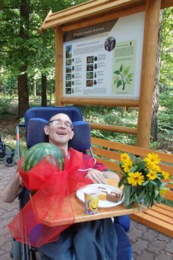 Mężczyzna na wózku inwalidzkim. Przed nim na stoliku leży arbóz, kwiaty i tależyk ciastem. W tle tablica informacyjna i drzewa.