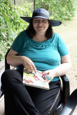 Kobieta w kapeluszu siedząca na krześle trzyma w rękach książkę i flamastry.