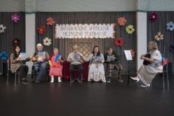 Siedem osób występuje na dużej sali. Za nimi udekorowana kwiatami ściana i napis Integracyjne Spotkanie Teatralno-Muzyczne.