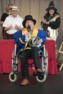 Mężczyzna na wózku inwalidzkim z mikrofonem w ręku. Za nim w tle stoją trzy inne osoby.