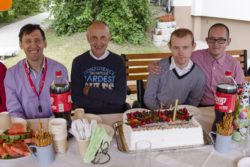 Czterej mężczyźni pozują do zdjęcia. Przed nimi stół na którym stoi tort i inne słodycze.