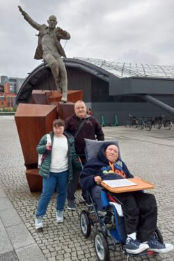 Kobieta i dwóch mężczyzn jeden z nich na wózku inwalidzkim, przed rzeźbą biegnącego mężczyzny.