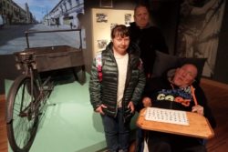 Kobieta i dwóch mężczyzn jeden z nich na wózku inwalidzkim, przed muzealną wystawą.