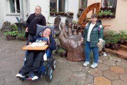 Kobieta i dwóch mężczyzn jeden z nich na wózku inwalidzkim, przed rzeźbą dużej dłoni.