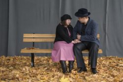 Na sali pełnej żółtych liści, na ławce siedzą trzymając się za ręce kobieta z mężczyną.