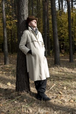 Elegancko ubrany mężczyzna z dłońmi schowanymi w kieszenie płaszcza stoi oparty o drzewo.