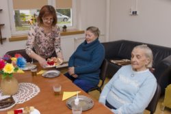 Kobieta nakłada ciasto siedzącym przy stole dwóm kobietom.