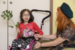 Dziewczynka na wózku inwalidzkim z różą w ręku i obok niej kobieta w zielonym berecie.