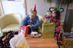 Siedzący w fotelu chłopiec wyjmuje prezent z papierowej torby. Obok niego na wózku inwalidzkim siedzi dziewczynka.