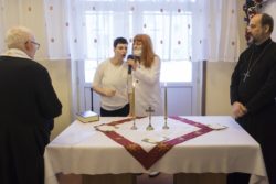 Dwie kobiety stojące przy stole na któym stoią krzyż, świece i leżą opłatki. Po bokach stołu stoi dwóch duchownych.