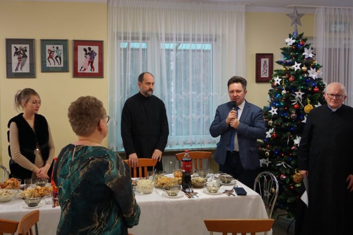 Przy świątecznie nakrytym stole stoją mężczyzna trzymający w dłoniach mikrofon, dwuch duchownych i dwie kobiety.