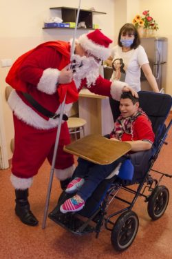 Mikołaj stojący przy siedzącej na wózku inwalidzkim dziewczynce. W tle stojąca kobieta.