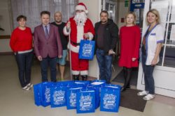 Zdjęcie grupowe sześciu osób z Mikołajem. Przed osobami na podłodze stoją torby z prezentami.