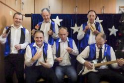 Sześciu mężczyzn w białych koszulach i niebieskich kamizelkach z instrumentami muzycznymi w rękach.