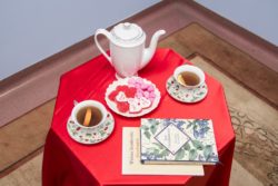 Stolik nakryty czerwonym obrusem na którym stoją filiżanki i dzbanek z herbatą, tależyk ze słodyczami i książki.