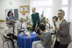 Siedem siedzących przy zastawionym słodyczami stoliku kobiet trzymających w dłoniach tulipany. Pomiędzy kobietami stoi mężcyna