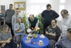 Cztery siedzące przy stoliku kobiety z tulipanami w dłoniach. Za nimi stoi sześciu mężczyzn. Jeden z nich nalewa herbatę do filiżanek.