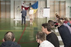 Na dużej sali stoi przed białą ławką mężczyzna i kobieta. Za nimi w tle dekoracja w barwach Polski i Ukrainy. o prawej stronie grupa siedzących osób.