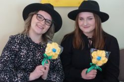 Dwie kobiety w czarnych kapeluszach z zółtymi kwiatkami w dłoniach.