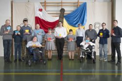 Zdjęcie grupowe. Kobiety i mężczyźni trzymające w rękach serca stoją na tle dekoracji w barwach Polski i Ukrainy.